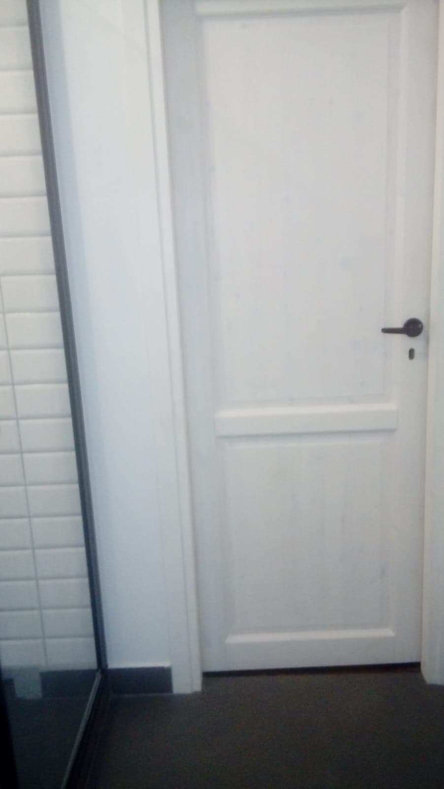 Białe, dwupanelowe drzwi wewnętrzne z czarną klamką, widoczne z bliska obok fragmentu ściany z białymi płytkami.