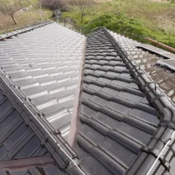 Widok z góry na fragment dachu pokrytego ciemną dachówką ceramiczną z zamontowanymi panelami słonecznymi i systemem odgromowym, na tle zieleni wzgórz.