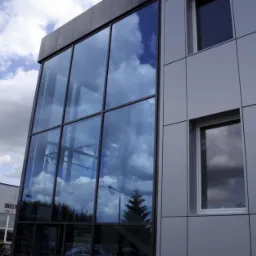 Nowoczesny budynek biurowy z elewacją z płyt warstwowych i dużymi oknami, w których odbija się błękitne niebo z chmurami.