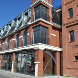 Odrestaurowany budynek z czerwonej cegły z metalowymi balustradami balkonowymi i przeszklonymi łukami na parterze, data 1892 na fasadzie.