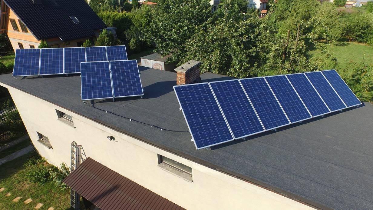 Instalacja paneli słonecznych na płaskim dachu budynku z widocznym kominem i drabina oparta o ścianę, otoczona zielenią.