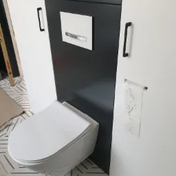 Podwieszana toaleta z zabudową szafkową w kolorach bieli i grafitu, z czarnymi uchwytami, na podłodze wzorzyste płytki, widoczna poziomica i rolka papieru toaletowego z dziecięcymi rysunkami.