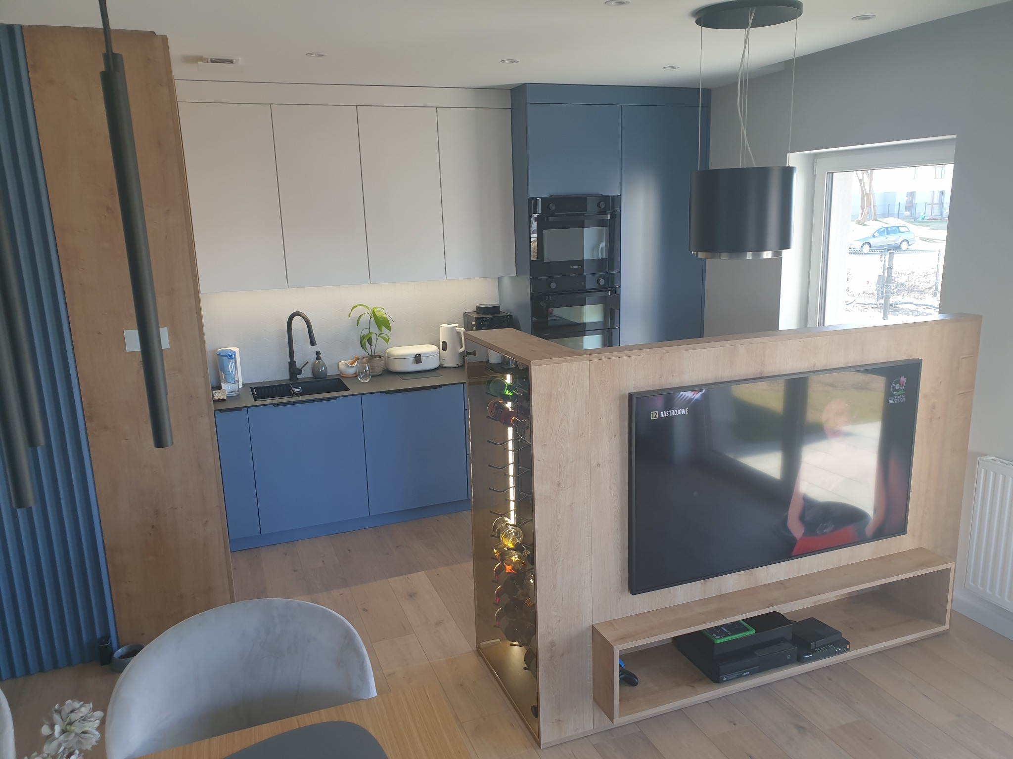 Nowoczesna kuchnia z niebieskimi i białymi szafkami, drewnianym blatem, zabudowaną ścianką działową z miejscem na telewizor i wbudowanym regałem na wino.