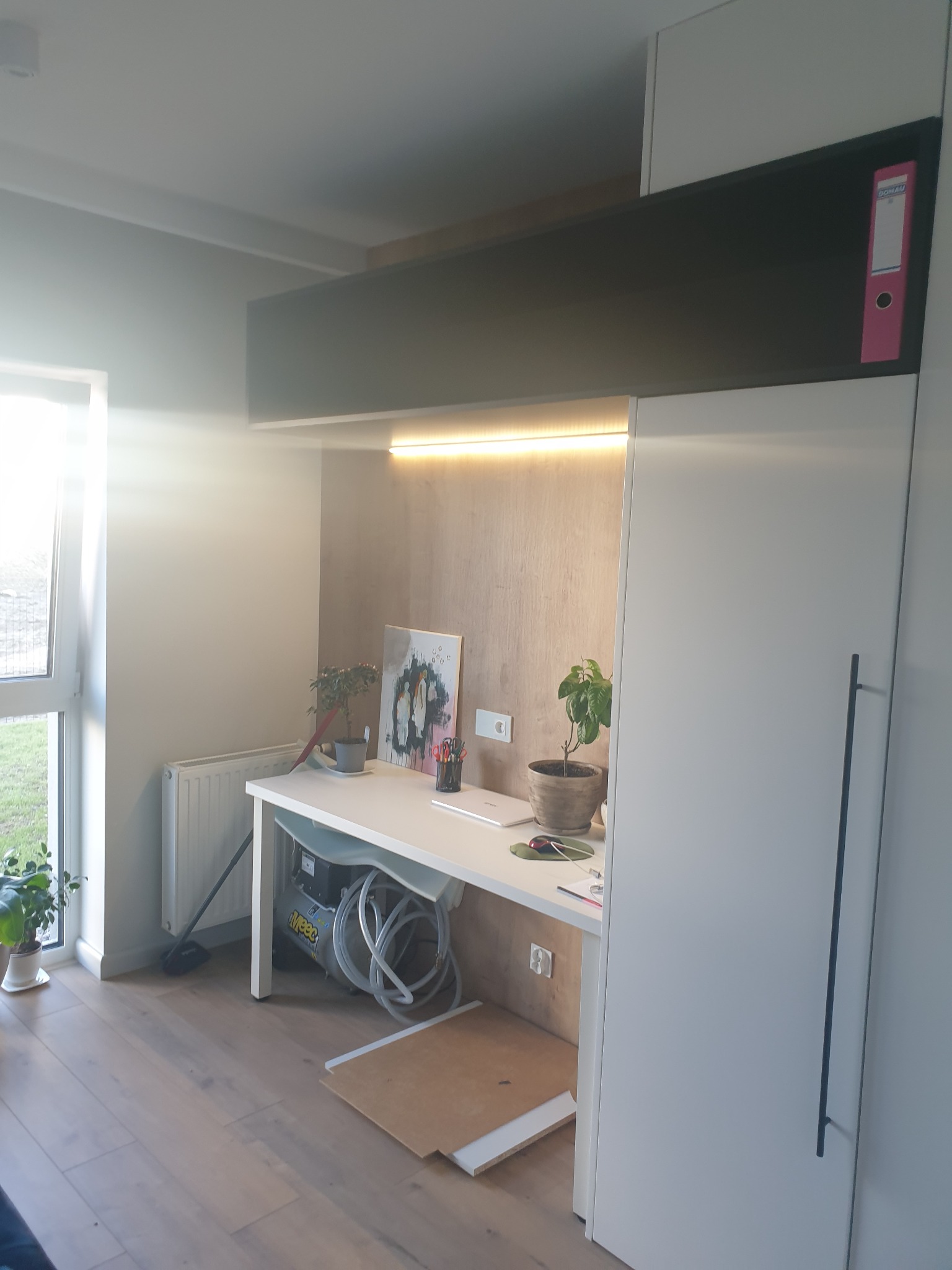 Biała szafa na wymiar z czarną nadstawką, biurko z roślinami i laptopem, oświetlenie LED, widoczne narzędzia montażowe na podłodze.