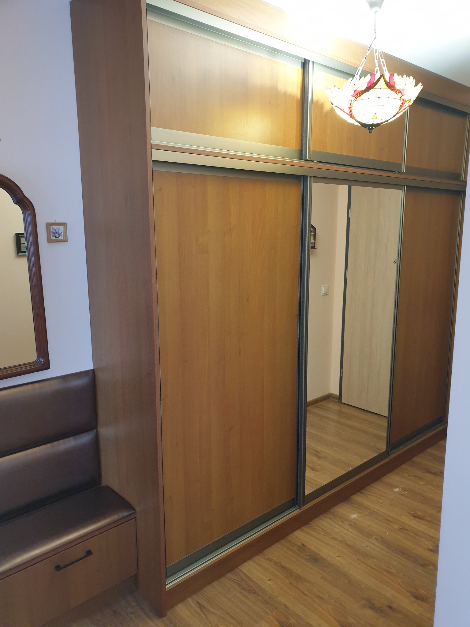 Szafa wnękowa z przesuwnymi drzwiami lustrzanymi, obok wbudowana ławka z szufladą, widoczny ozdobny żyrandol.