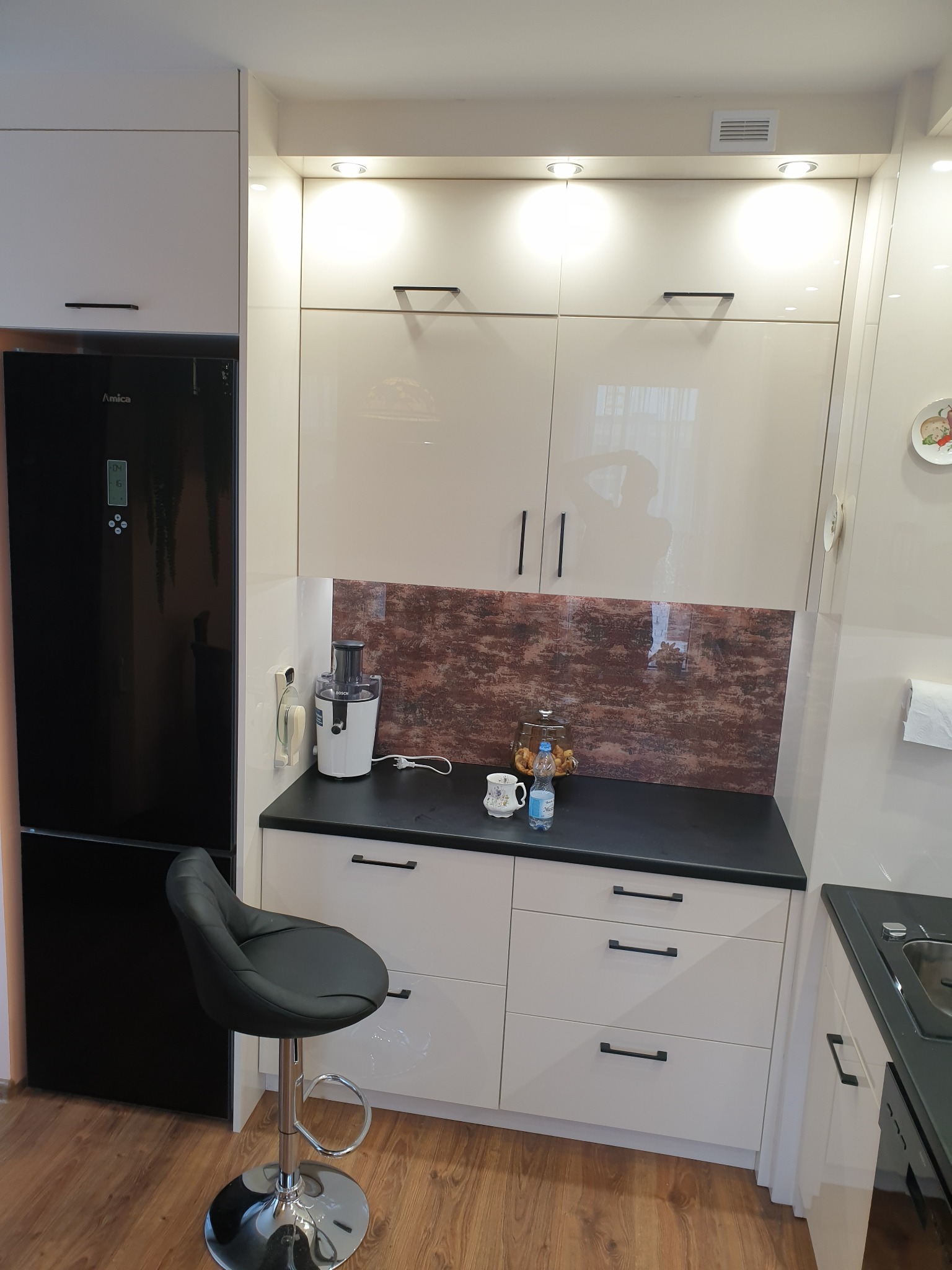 Jasna kuchnia z czarnym blatem i frontami szafek z podłużnymi uchwytami, czarna lodówka Amica i hoker przy blacie, na blacie wyciskarka do soków i słoik z ciastkami.