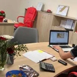 Mężczyzna w okularach siedzi przy biurku w biurze, tyłem do kamery, korzystając z laptopa. Na biurku kwiaty w wazonie, kalkulator, dokumenty i miseczka z przekąskami. W tle meble biurowe, czerwone...