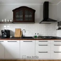 Nowoczesna kuchnia z białymi szafkami, drewnianym blatem, czarną armaturą i białymi płytkami na ścianie, uzupełniona o drewnianą szafkę wiszącą i minimalistyczny okap.