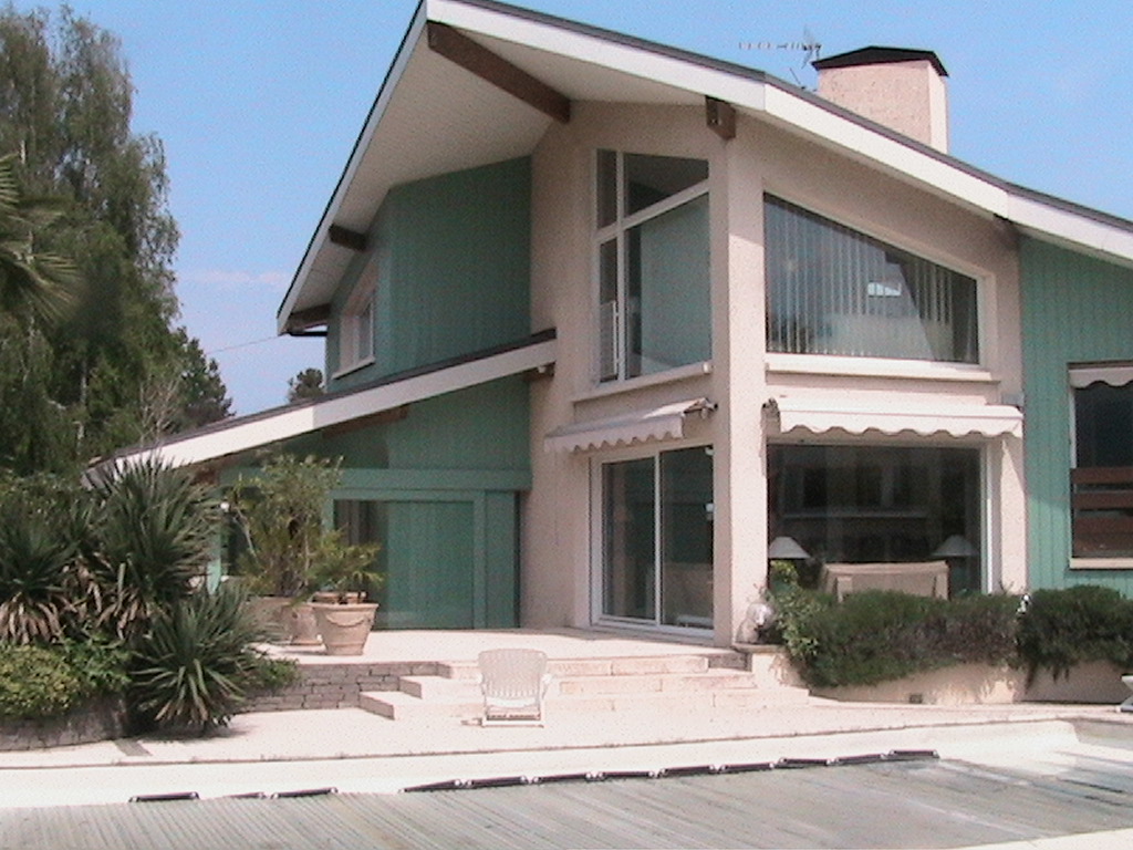 Dwukondygnacyjny dom z zieloną elewacją, dużymi oknami i schodami prowadzącymi do basenu, widoczne rośliny w donicach.