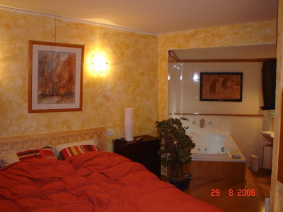 Sypialnia z łóżkiem z czerwonym nakryciem i ścianami w odcieniach żółci, z widokiem na łazienkę z wanną z hydromasażem za szklaną przegrodą.