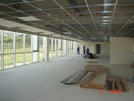 Wnętrze biurowe w trakcie remontu: widoczne metalowe profile sufitowe, częściowo zamontowane płyty, duże okna z widokiem na zieleń, oraz trzech pracowników w oddali, przygotowujących się...