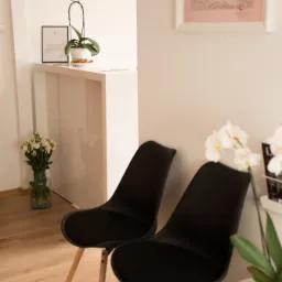 Jasne wnętrze poczekalni: dwa czarne krzesła z drewnianymi nogami, biały stolik z orchideą, wazon z białymi kwiatami, obraz na ścianie i minimalistyczne dekoracje.