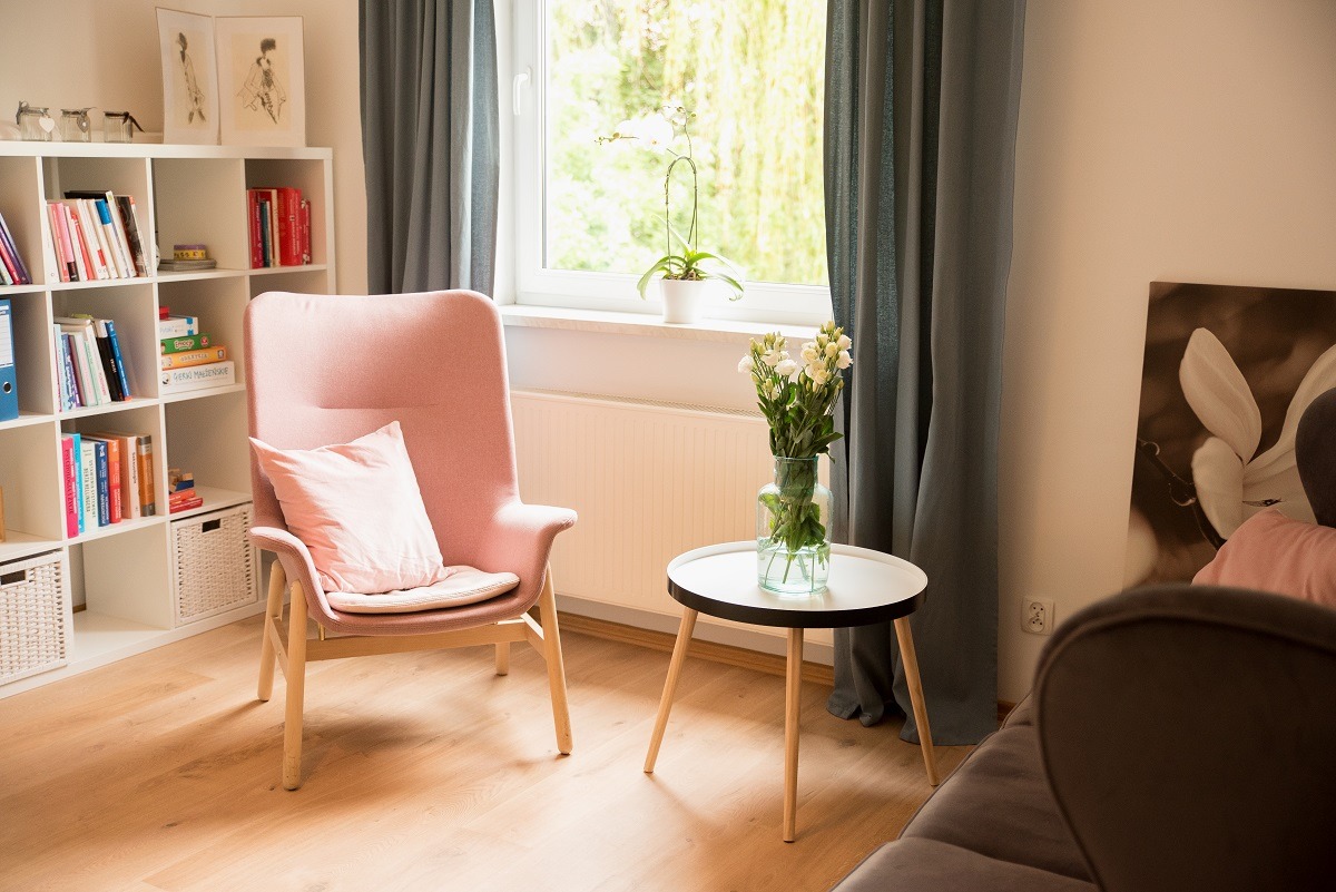 Jasny pokój z różowym fotelem, poduszką, białym regałem z książkami, kwiatami w wazonie na okrągłym stoliku i obrazem magnolii na ścianie.