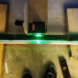 Instal-Pro, Krzysztof Kuś - Laser krzyżowy emituje zieloną wiązkę, wyznaczając poziom na ramie montażowej. Widoczne deski, poziomica i fragment obuwia. Ujęcie z góry, w trakcie prac remontowych.