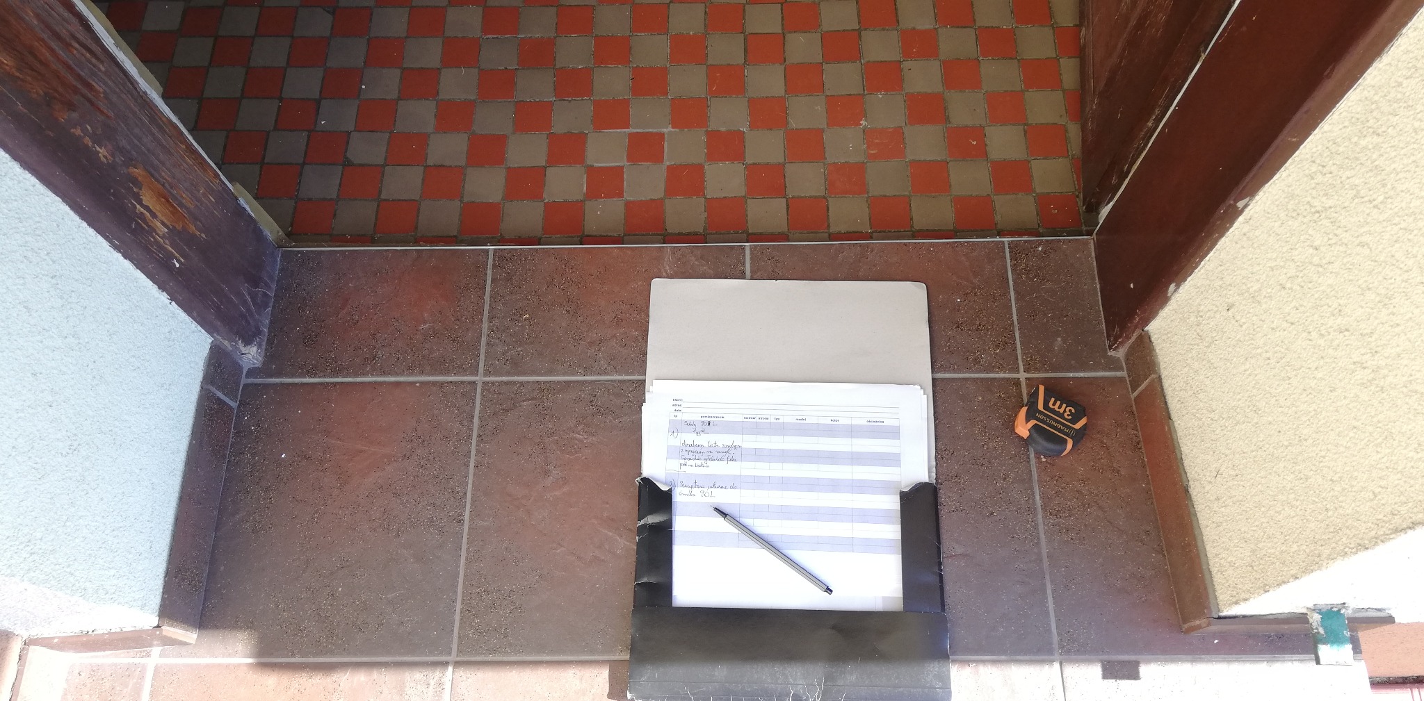 Widok z góry na próg drzwiowy z brązowymi płytkami, na którym leży otwarty segregator z dokumentami i ołówkiem, obok leży zwinięta miarka.