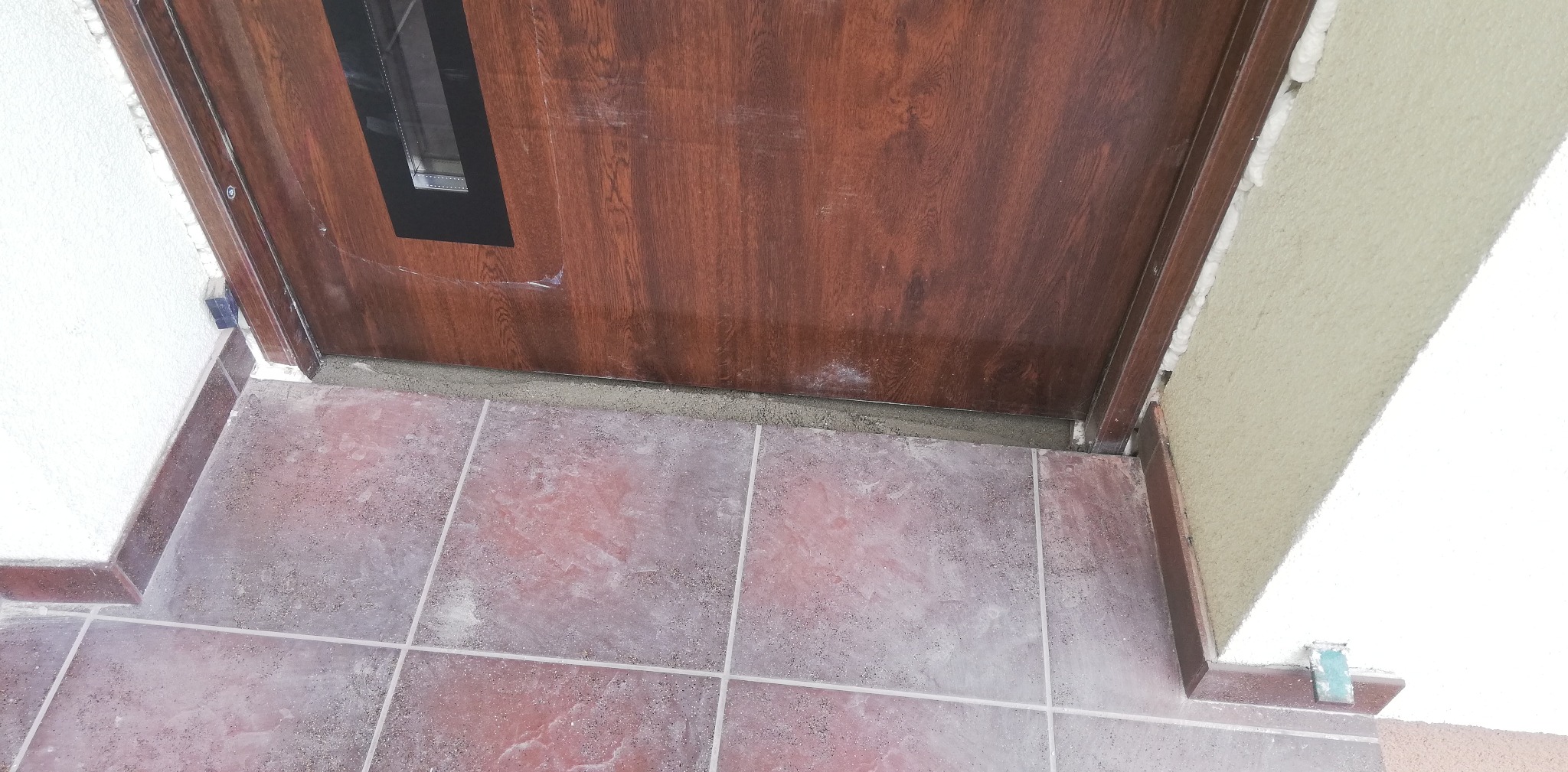 Brązowe drzwi wejściowe z czarnym, prostokątnym okienkiem, częściowo osadzone w ościeżnicy, z widoczną pianką montażową i szarą zaprawą u dołu, na tle brązowo-czerwonej podłogi z płytek.
