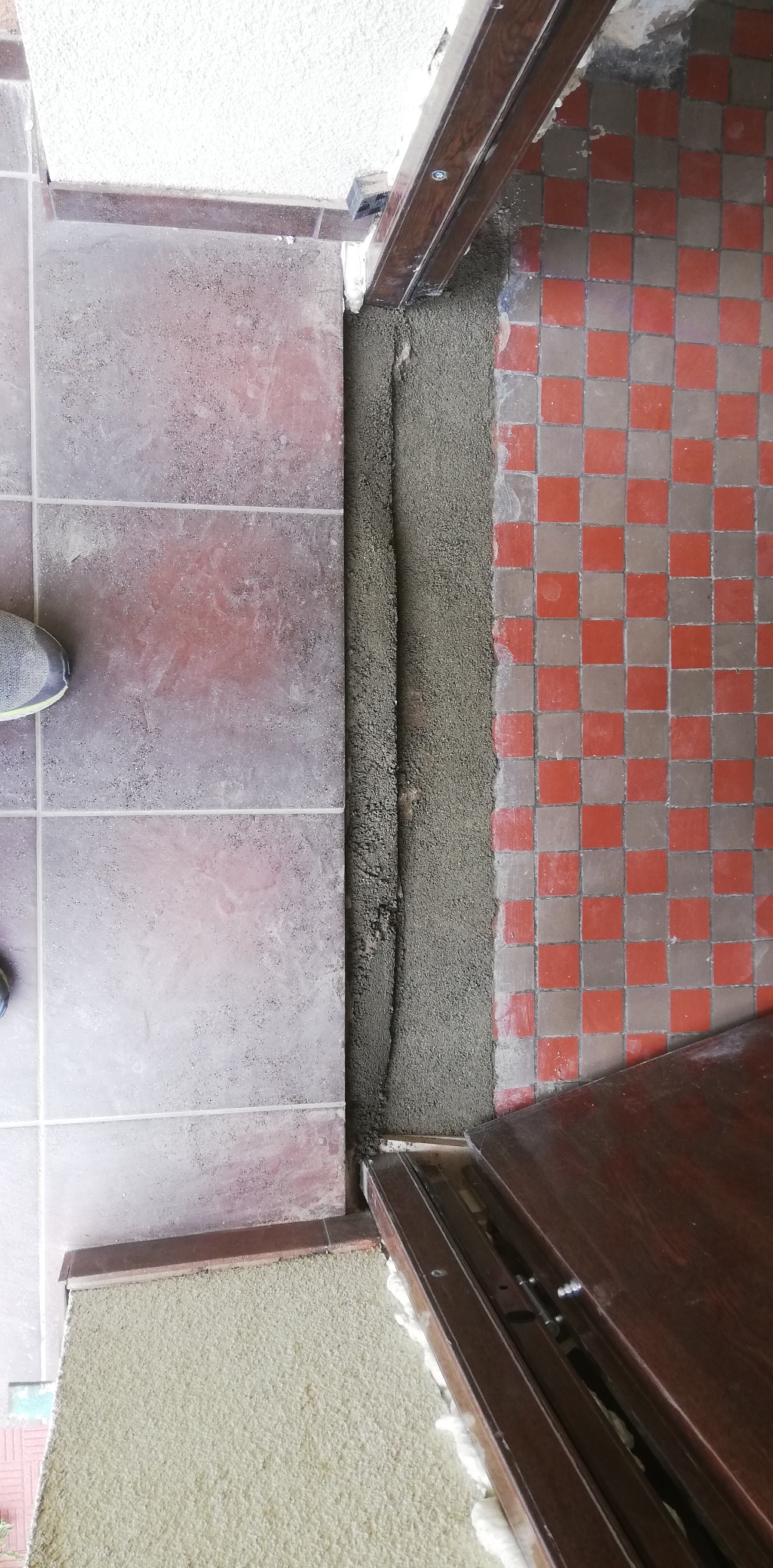 Widok z góry na próg w trakcie osadzania ościeżnicy, widoczne płytki podłogowe po obu stronach oraz wypełniona zaprawą szczelina między nimi. Pianka montażowa widoczna przy ościeżnicy.