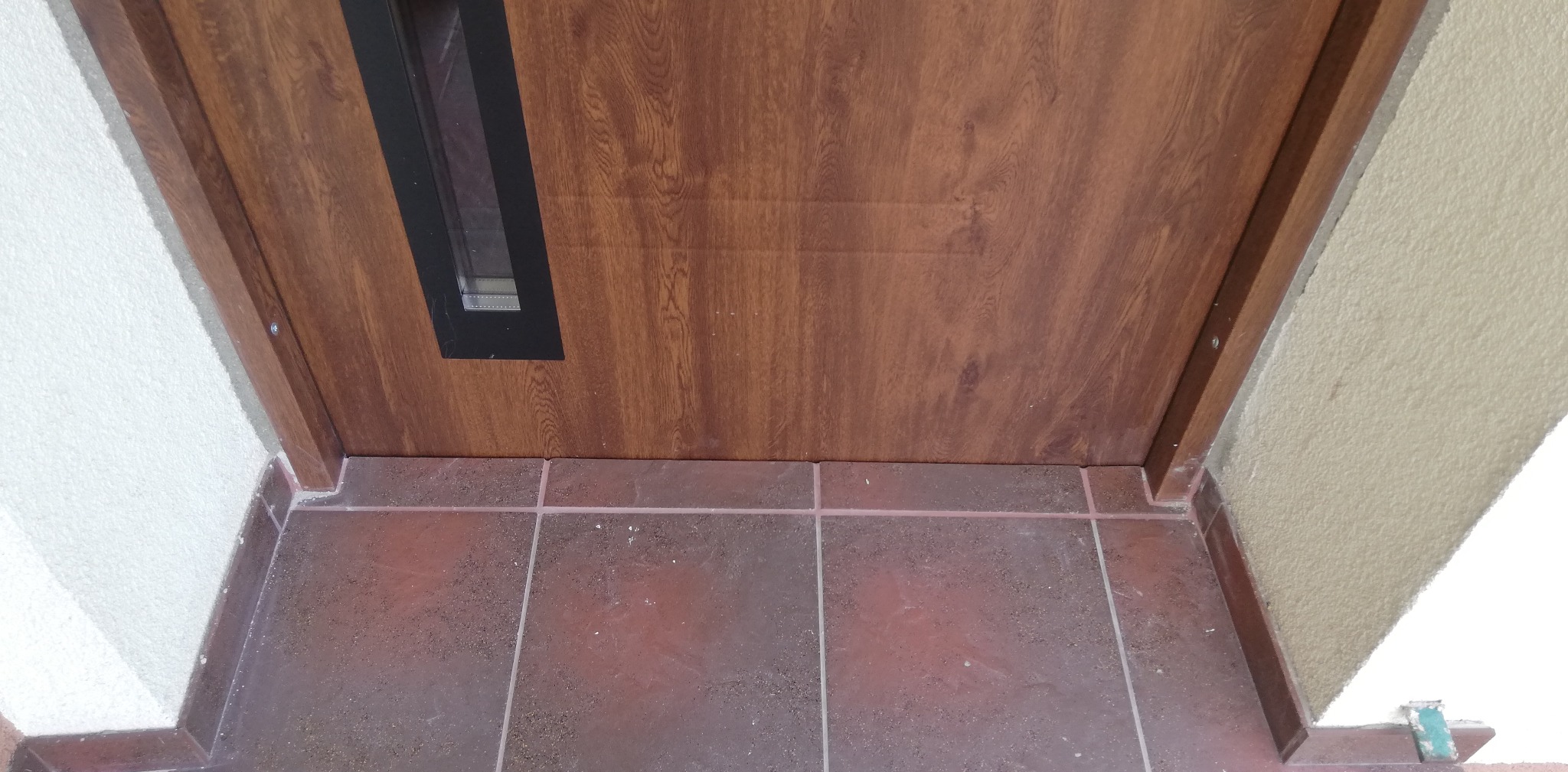 Fragment brązowych drzwi z małym okienkiem od dołu, widok na próg i brązowe płytki podłogowe oraz fragment ościeżnicy i ściany o nierównej fakturze.