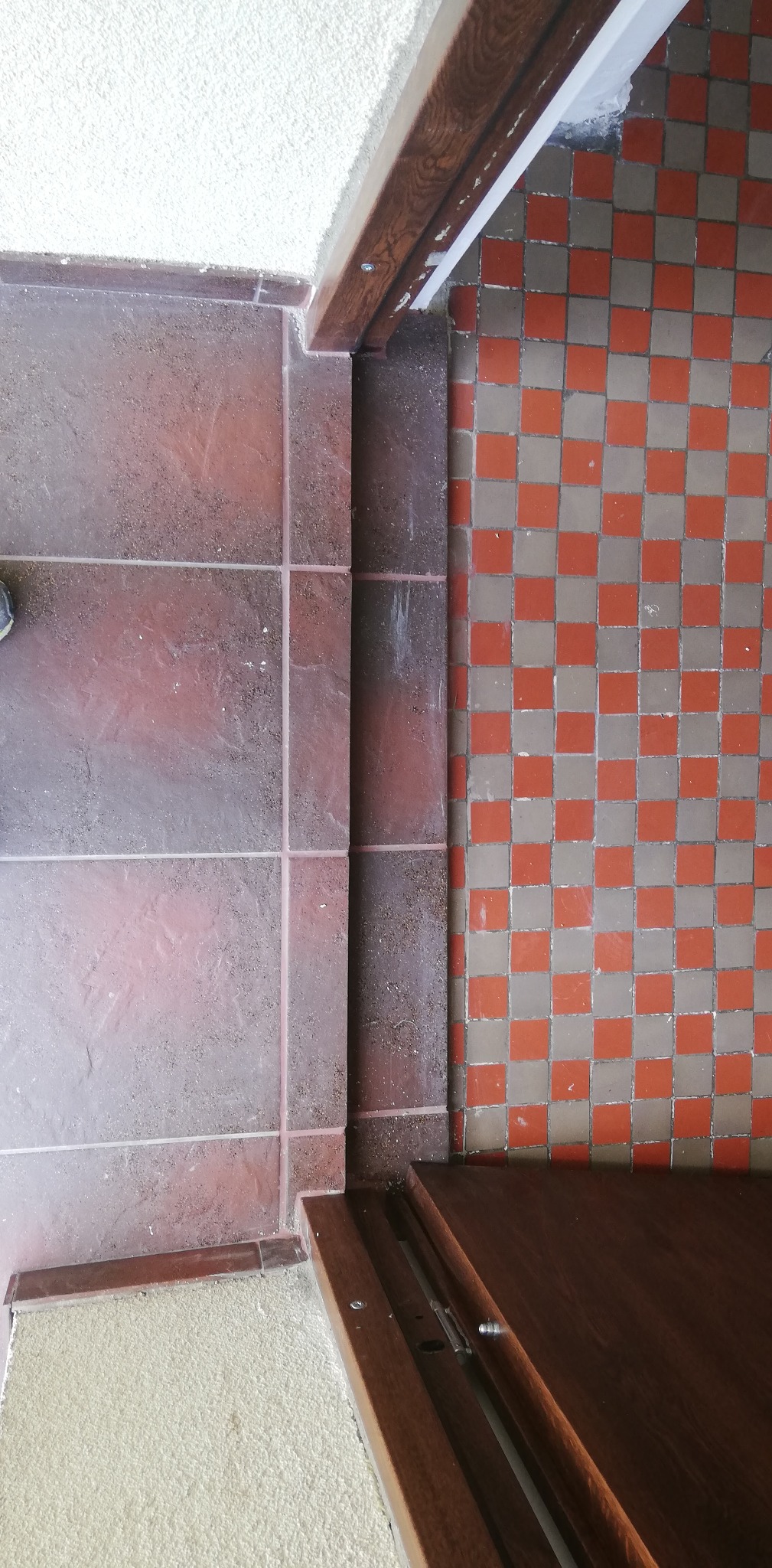 Fragment podłogi z brązowych płytek i szachownicy czerwono-szarych płytek, widoczna dolna część drewnianej futryny z zamontowanym mechanizmem, biały tynk na ścianie.