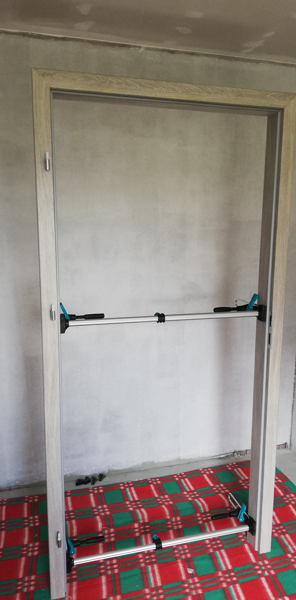 Ościeżnica drzwiowa z drewna w trakcie montażu, zabezpieczona rozpórkami. Widoczne narzędzia monterskie i surowa ściana w tle. Podłoga zabezpieczona kocem.