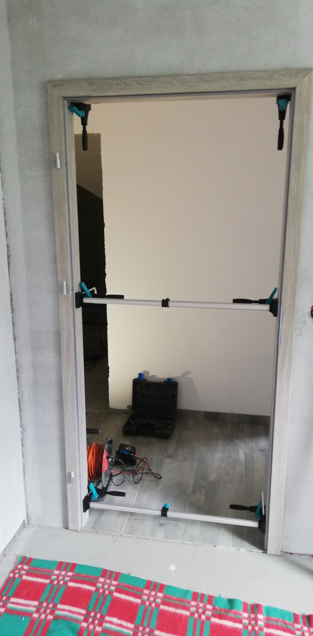 Montaż ościeżnicy drzwiowej z użyciem rozpórki teleskopowej i zacisków. Widoczna skrzynka narzędziowa i zwinięty przedłużacz. Ściany w surowym stanie.