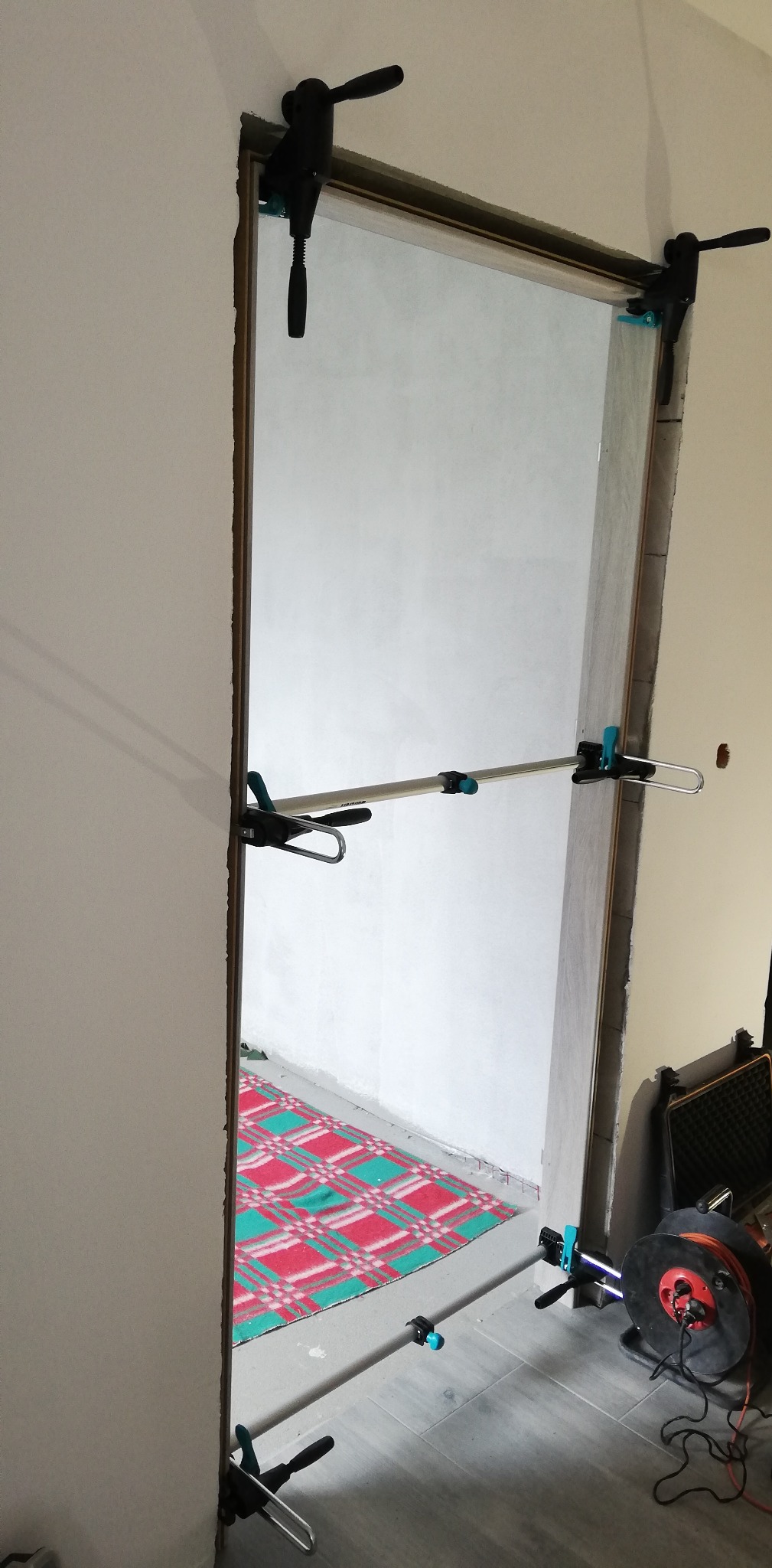 Montaż ościeżnicy drzwiowej z użyciem rozpórek i zacisków. Widoczny fragment podłogi z wzorzystym dywanem i zwinięty kabel. Ściany w kolorze białym i szarym.