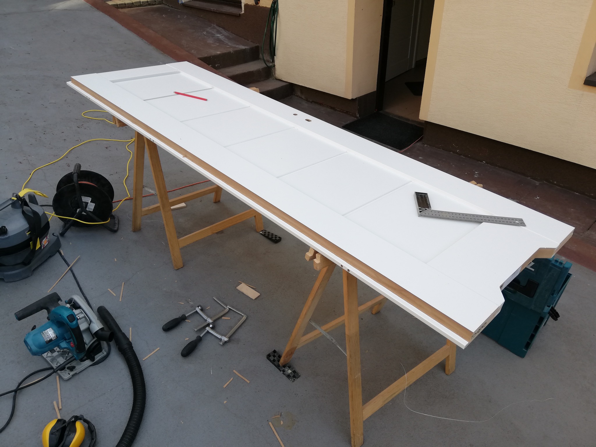 Białe drzwi panelowe na kozłach, w trakcie obróbki. Widoczne narzędzia: piła, kątownik, ściski. Przygotowanie do montażu, zarys ołówkiem. Detal pracy.