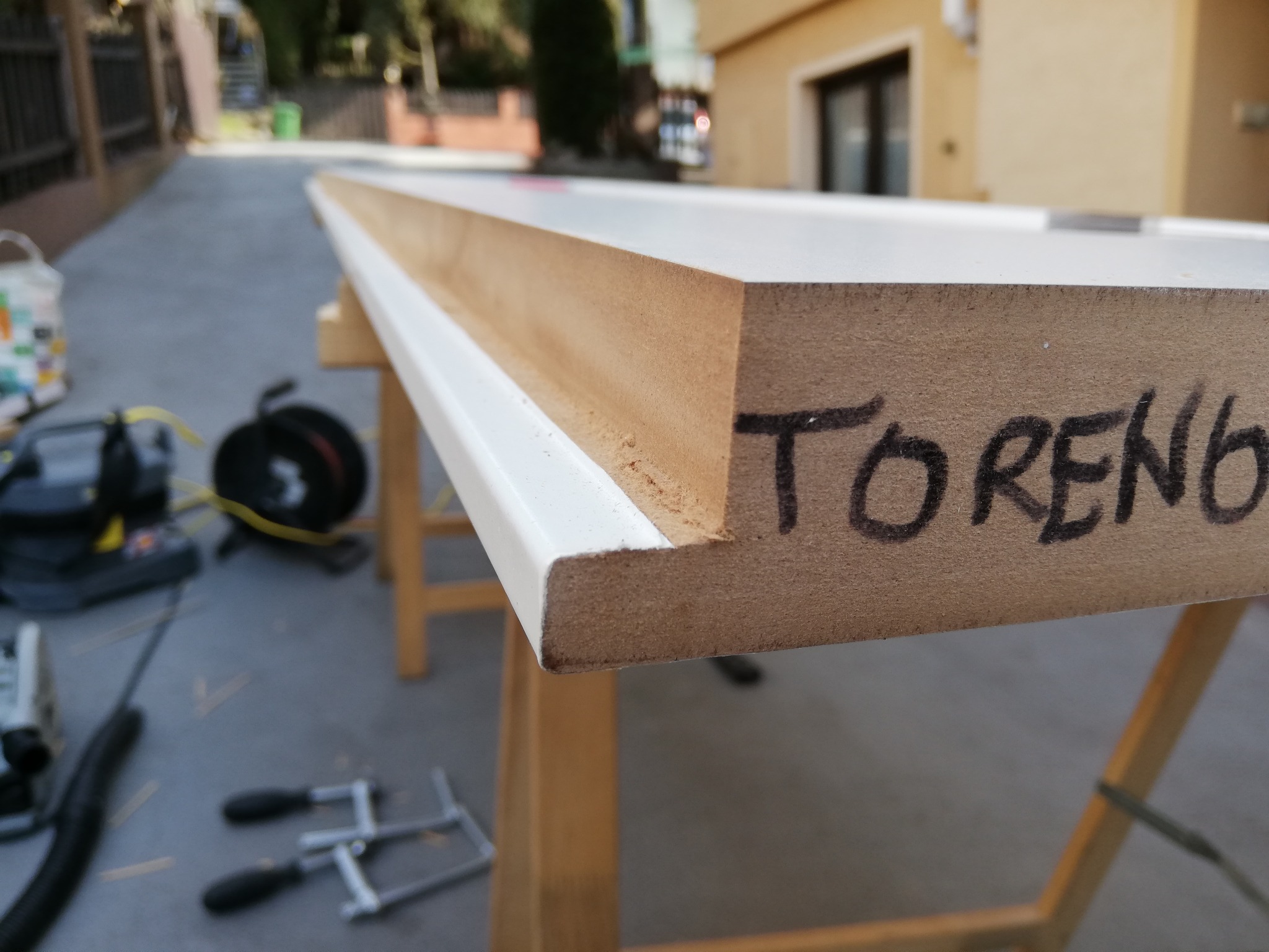 Fragment drzwi z napisem 'TORENB' na drewnianej powierzchni, leżący na kozłach. W tle narzędzia i otoczenie zewnętrzne.