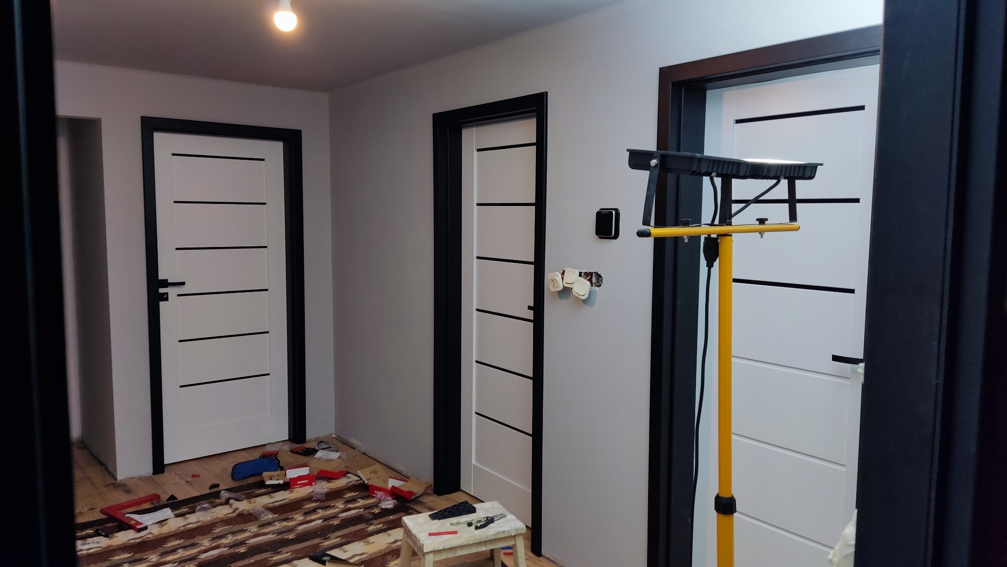 Montaż drzwi wewnętrznych z czarną ościeżnicą i białym skrzydłem z poziomymi frezami. Na podłodze widoczne ślady po montażu, oświetlenie budowlane.