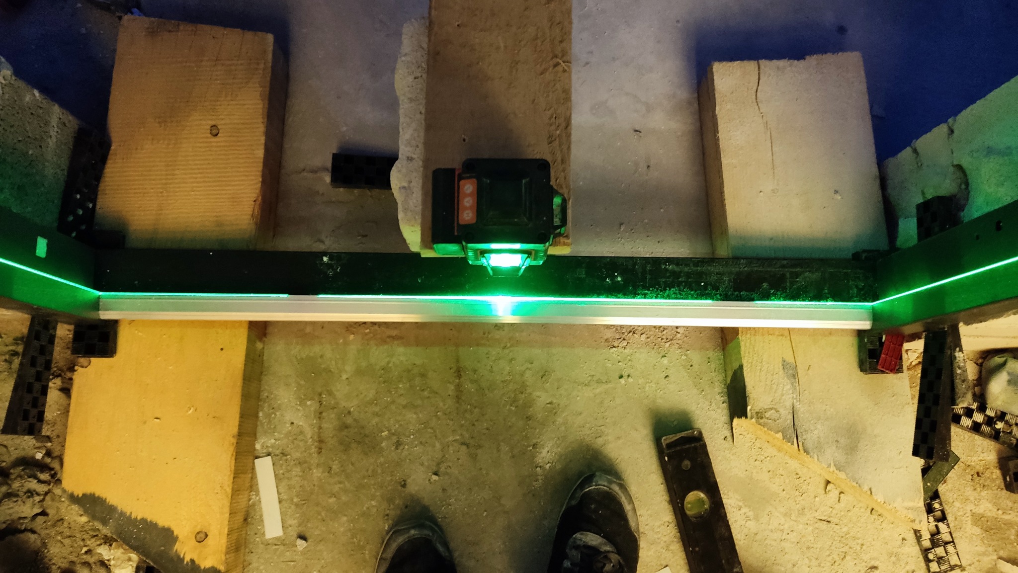 Laser krzyżowy emituje zieloną wiązkę, wyznaczając poziom na ramie montażowej. Widoczne deski, poziomica i fragment obuwia. Ujęcie z góry, w trakcie prac remontowych.