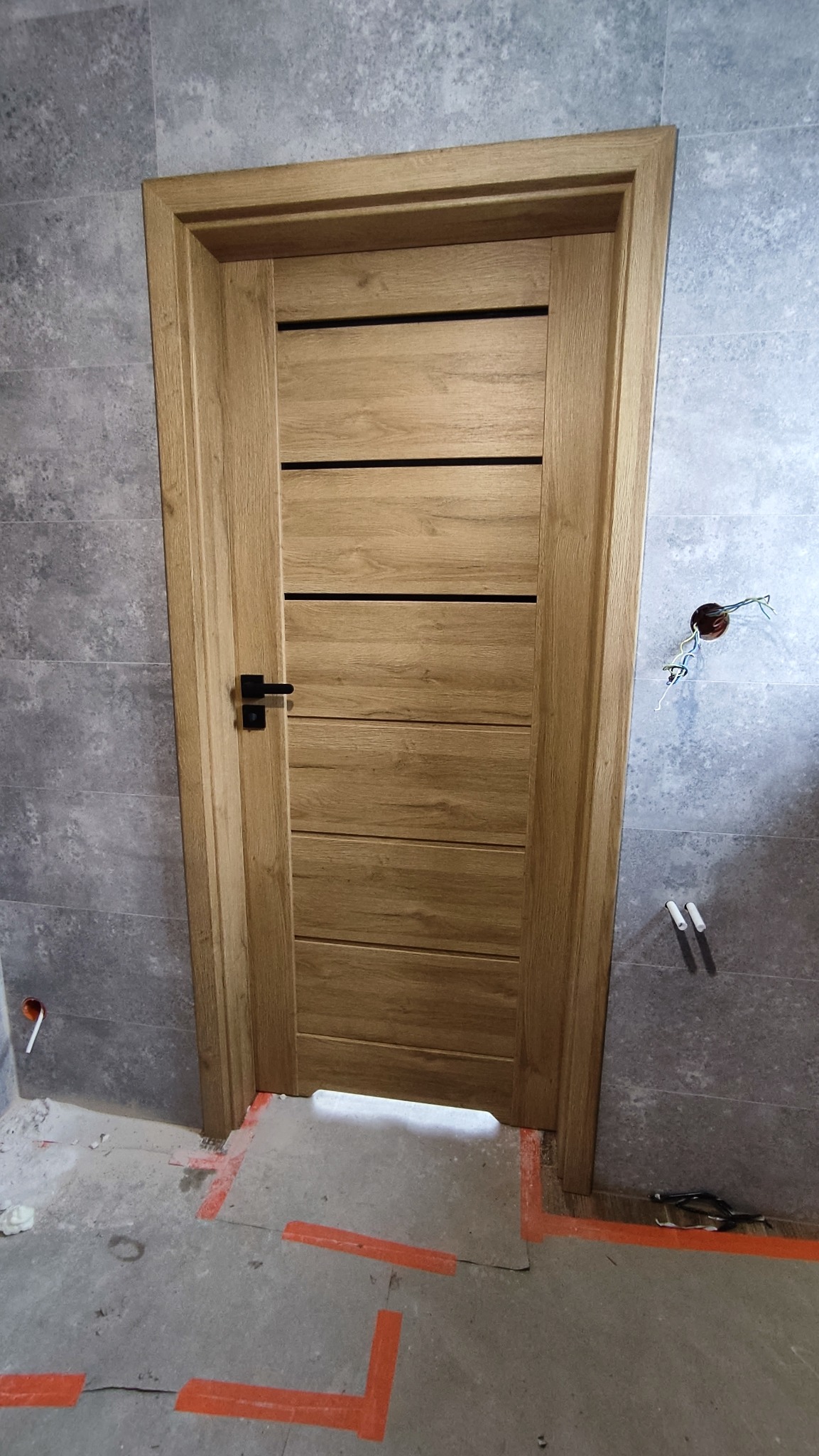 Dębowa, nowoczesna stolarka drzwiowa z czarną klamką, zamontowana w łazience z szarą, kamienną okładziną. Widoczne ślady po remoncie.