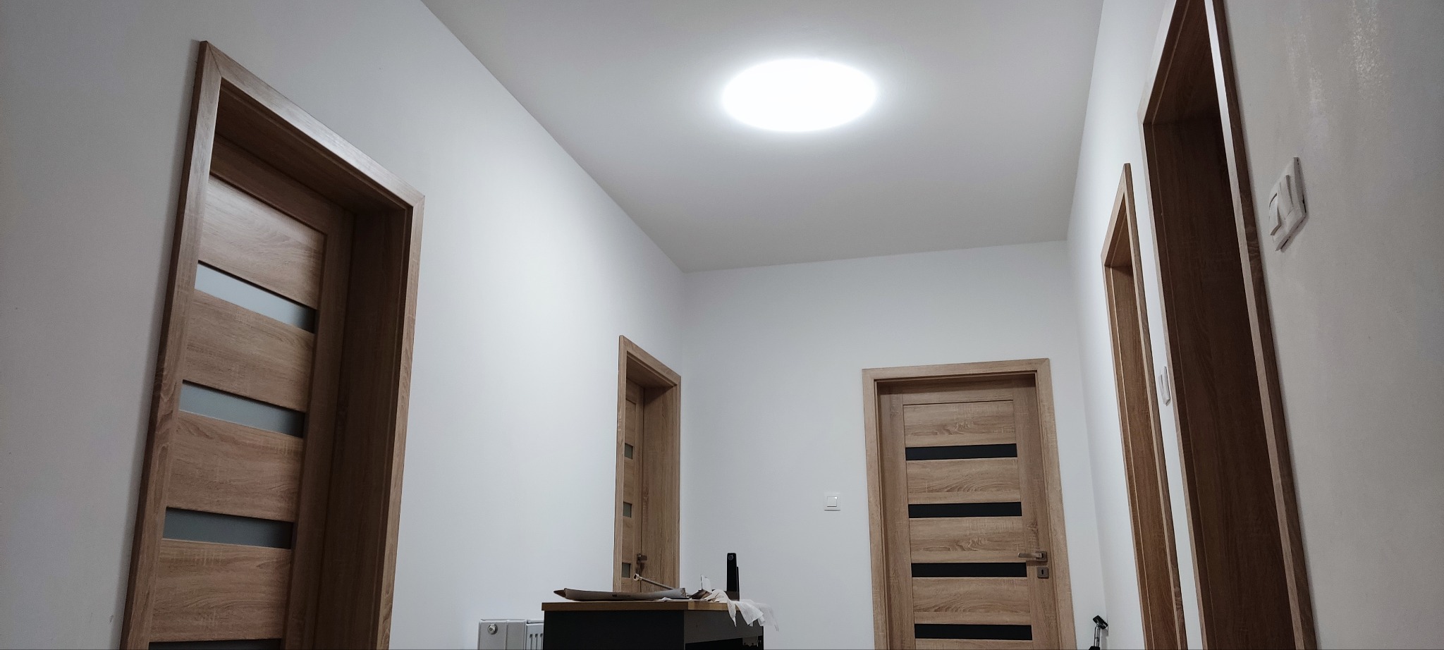 Jasny korytarz z trzema drewnianymi drzwiami z przeszkleniami, oświetlony okrągłym świetlikiem w suficie. Minimalistyczny design, białe ściany, widoczny fragment mebla.