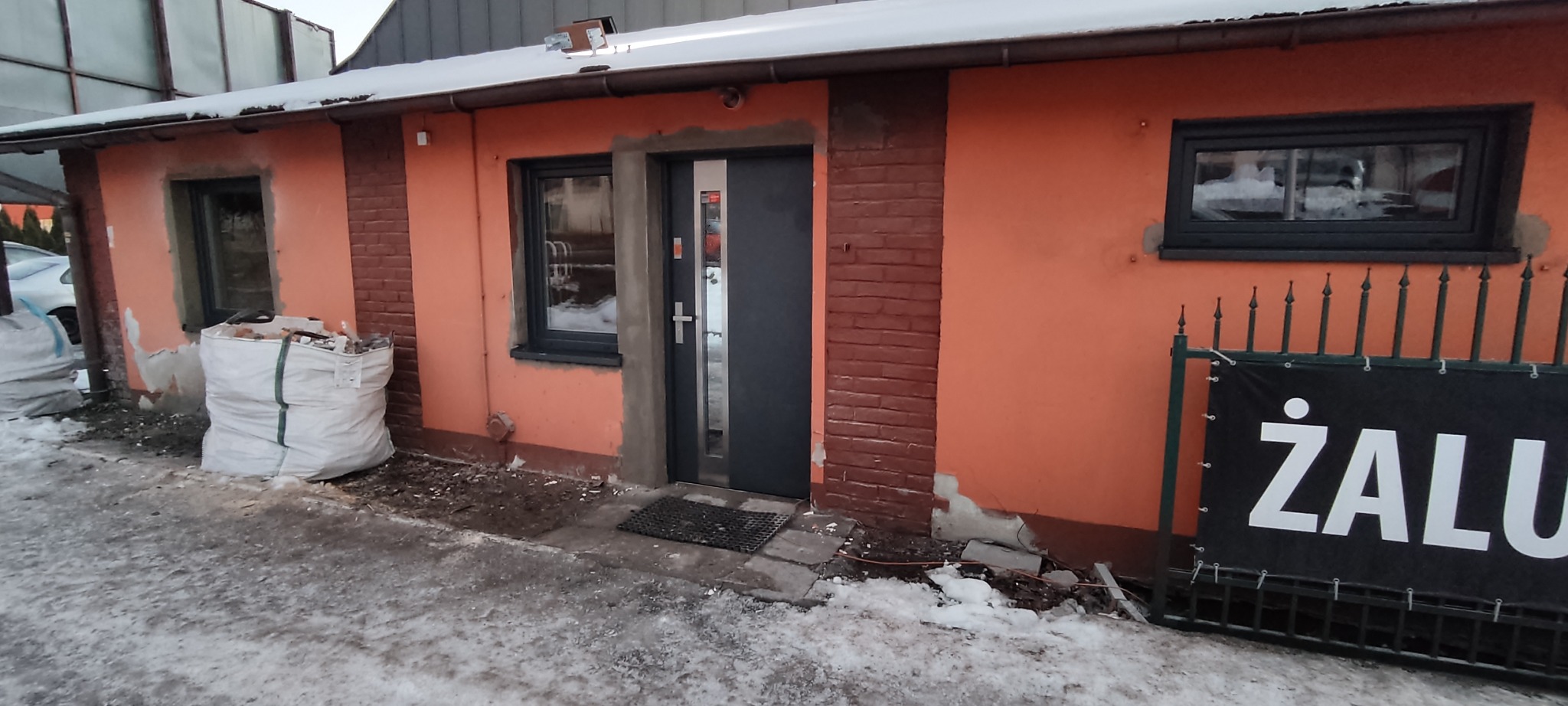 Budynek z pomarańczową elewacją po montażu okien i drzwi. Widoczne ślady prac murarskich i worki z gruzem. Zima.