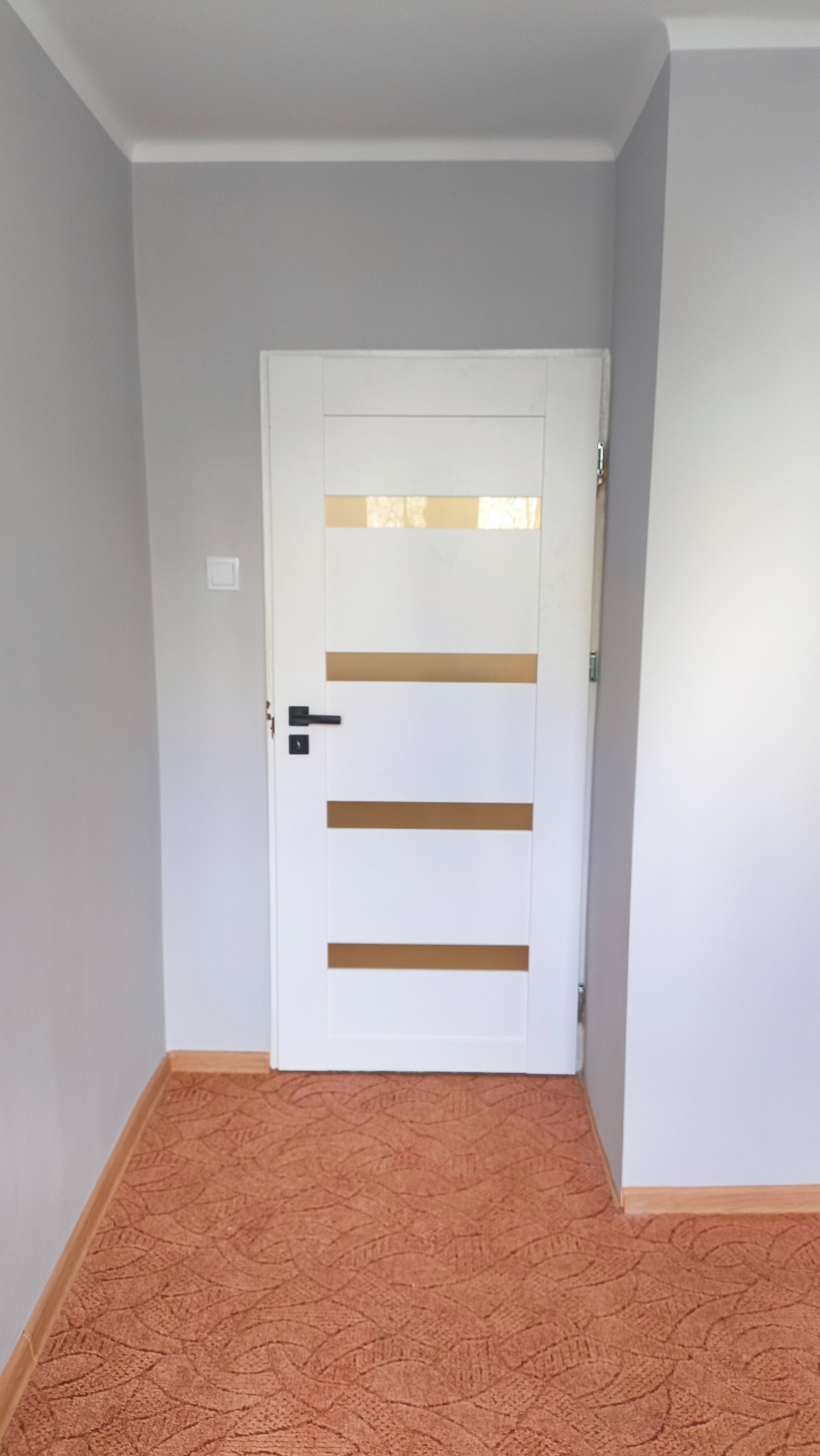 Białe drzwi wewnętrzne z poziomymi, złotymi wstawkami, czarna klamka, szare ściany i brązowa wykładzina podłogowa. Widok na zamontowane drzwi w korytarzu.
