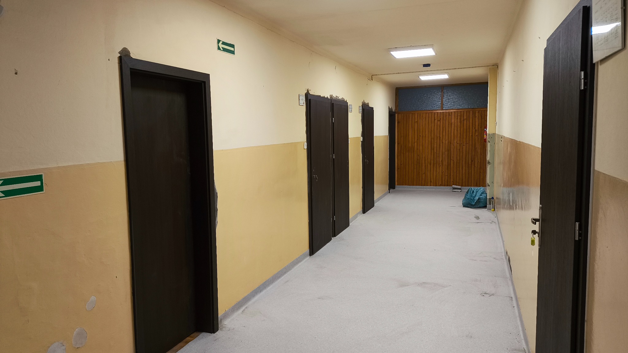 Korytarz po montażu ciemnych drzwi w Cieszynie. Widoczne ślady po pracach wykończeniowych na ścianach i podłodze. Strzałki ewakuacyjne na ścianie.