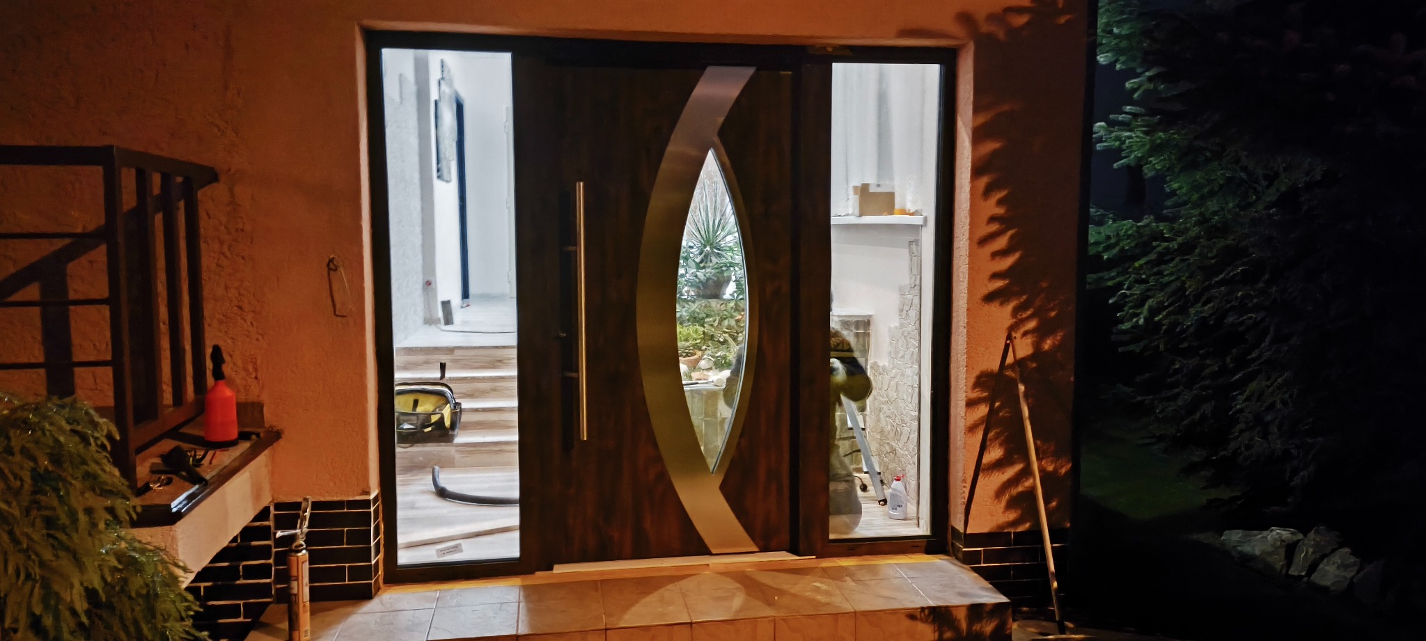 Nowoczesne, brązowe drzwi wejściowe z przeszkleniem w kształcie łzy, widoczne narzędzia i schody wewnątrz. Oświetlenie nocne, ciemne tło z zielenią.