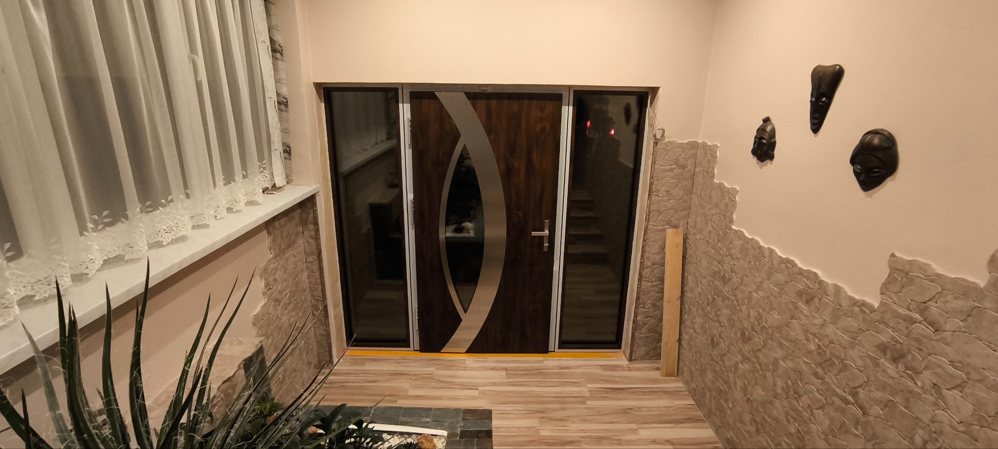 Nowoczesne drzwi wejściowe z przeszkleniami bocznymi, brązowe z srebrnym, dekoracyjnym pasem. Widoczne elementy dekoracyjne na ścianie i roślina doniczkowa.