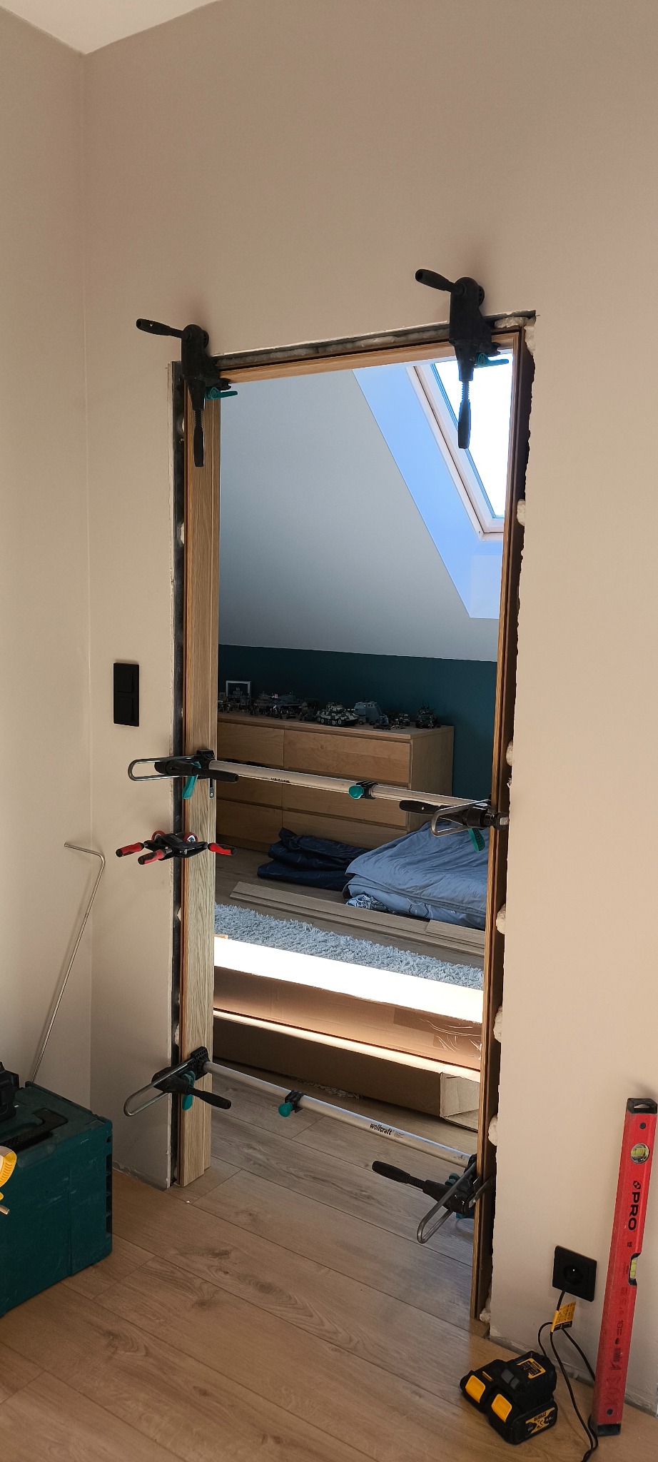 Montaż ościeżnicy drzwiowej z użyciem rozpór, widoczne narzędzia i poziomica. W tle pokój z oknem dachowym i komodą z zabawkami.