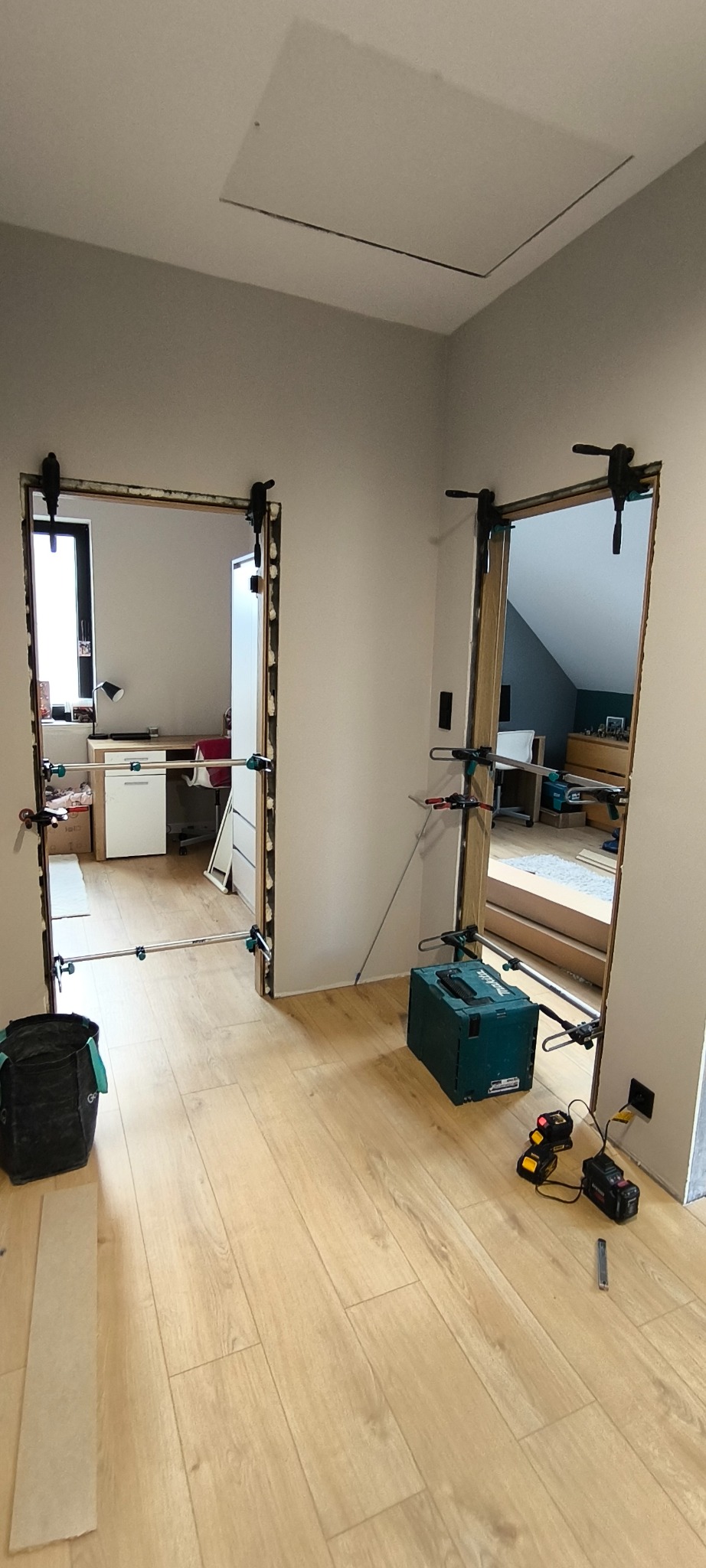 Przygotowanie do montażu dwóch ościeżnic w jasnym wnętrzu. Widoczne narzędzia, poziomice i klamry mocujące. W tle widoczne pomieszczenia z wyposażeniem.