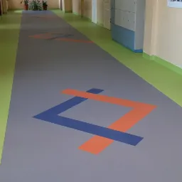 Długi korytarz z szarą wykładziną podłogową ozdobioną powtarzalnym wzorem geometrycznym w kolorach pomarańczowym i niebieskim, z zielonymi listwami przypodłogowymi i beżowymi ścianami.