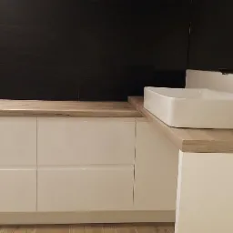 Biała szafka łazienkowa z drewnianym blatem, na którym stoi prostokątna umywalka i czarna bateria, na tle czarnych płytek.