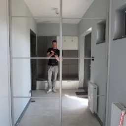 Dwudrzwiowa szafa przesuwna z lustrzanymi frontami, odbijająca wnętrze przedpokoju z siedziskiem i wieszakami, fotografowana z perspektywy osoby robiącej zdjęcie w lustrze.