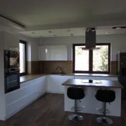 Nowoczesna, jasna kuchnia z białymi frontami szafek, drewnianym blatem i wyspą z dwoma czarnymi hokerami na chromowanych nogach; widok na ogród przez okno.