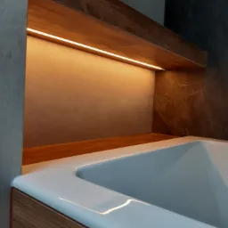 Biała wanna obudowana drewnopodobną płytą, z podświetlaną półką nad nią, oświetlenie LED w ciepłej barwie, szare płytki na ścianach.