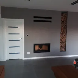 Nowoczesny salon z szarymi ścianami, wbudowanym kominkiem z paleniskiem i niszą wypełnioną drewnem opałowym obok, białe drzwi z poziomymi przeszkleniami, ciemna podłoga.