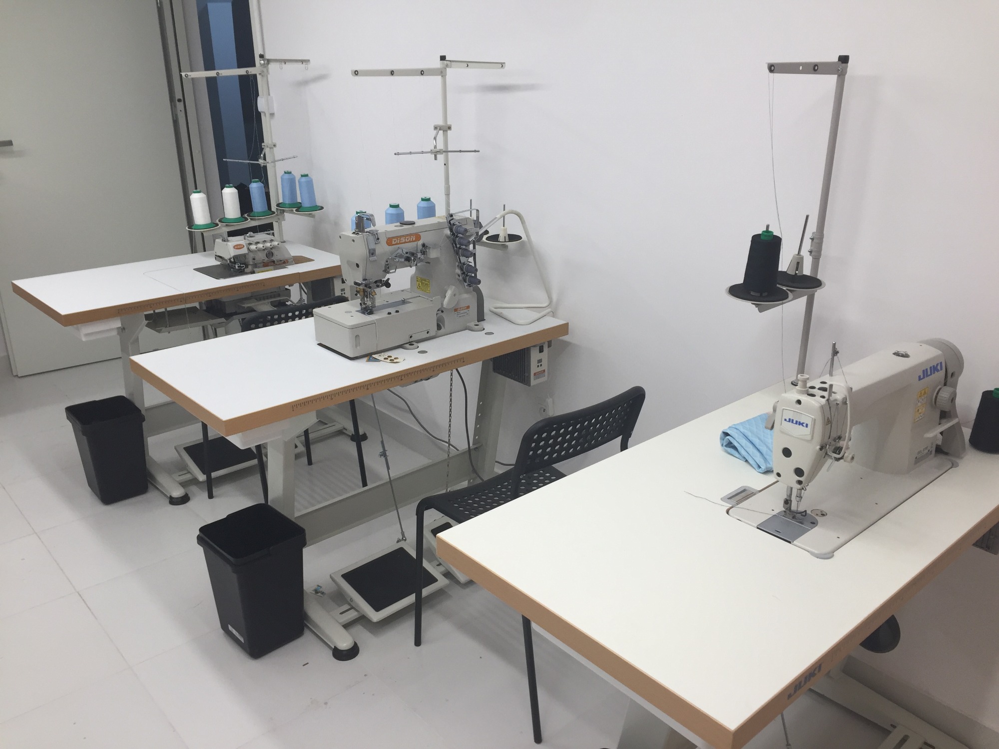 Trzy stanowiska pracy z przemysłowymi maszynami do szycia, każda z białym blatem i pedałem nożnym, z widocznymi szpulkami nici i czarnymi koszami na ścinki.