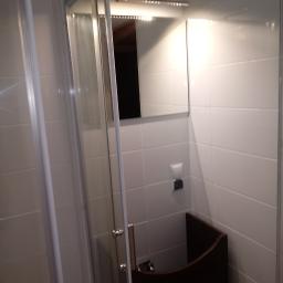 Celdom Spec - Wnętrze łazienki z kabiną prysznicową z przesuwnymi drzwiami, lustrem z oświetleniem LED i ciemną szafką pod umywalką, wykończone białymi płytkami.