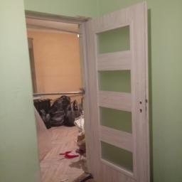 Celdom Spec - Otwarta, biała, drewnopodobna ościeżnica z czterema przeszkleniami, widok na remontowane wnętrze z workami gruzu i narzędziami, ściany w odcieniach zieleni i beżu.