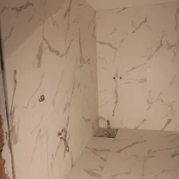 Białe płytki imitujące marmur na ścianach i podłodze w trakcie montażu, widoczne przyłącza wodne i otwory na gniazdka, ślady zaprawy na krawędzi ściany.