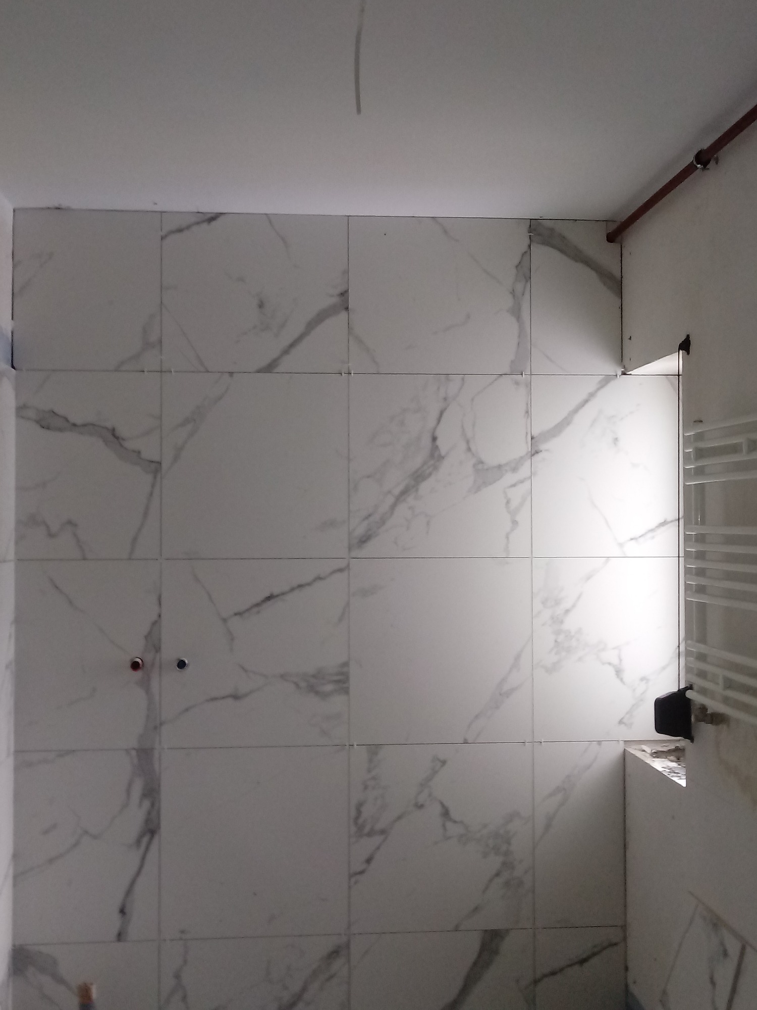Ściana łazienki wyłożona białymi płytkami imitującymi marmur, widoczne krzyżyki dystansowe i wystające rury instalacji wodnej